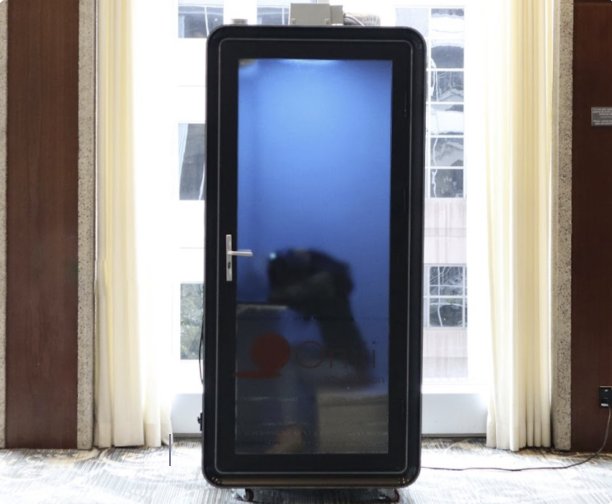 Open Booth, a black soundproof pod with a glass door, in a bright room